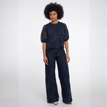 Load image into Gallery viewer, A model with an afro hairstyle wearing a matching navy blue two-piece set consisting of a short-sleeve cropped top with sheer puff sleeves and matching wide-leg trousers, both featuring an intricate floral eyelet lace pattern. She is standing against a plain white studio background.
