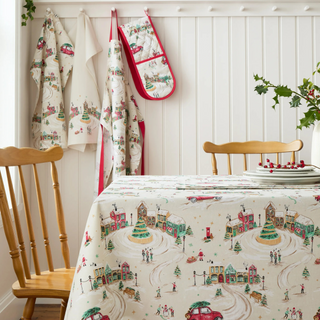 Christmas-themed tablecloth with village design on a table with wooden chairs.