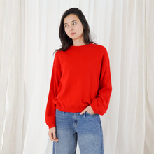 Load image into Gallery viewer, A woman wearing a bright red crewneck knit sweater and blue jeans, standing in front of a white curtain backdrop.
