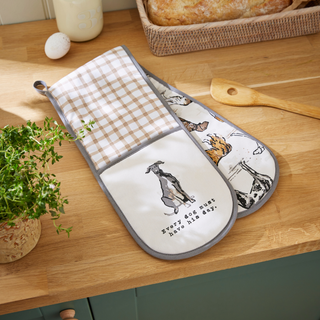 Set of two oven mitts on a wooden surface with a plant and bread basket in the background.