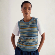 Load image into Gallery viewer, A woman wearing a blue and navy patterned knit sweater vest over a white t-shirt and dark blue pants, standing against a plain off-white wall.
