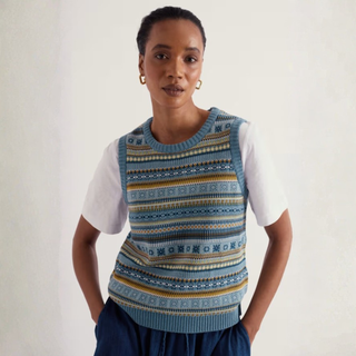 A woman wearing a blue and navy patterned knit sweater vest over a white t-shirt and dark blue pants, standing against a plain off-white wall.