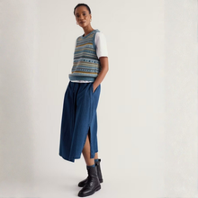 Load image into Gallery viewer, A model stands against a light off-white studio background wearing a sleeveless knit vest with blue, yellow, and gray geometric patterns over a white t-shirt, paired with a long blue pleated skirt and black ankle boots.
