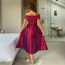 Load image into Gallery viewer, Woman in a burgundy off-shoulder dress standing in a bedroom.
