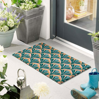 Patterned doormat on a floor with potted plants and shoes in the background