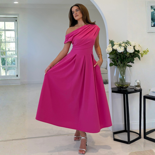 Woman in a pink one-shoulder dress standing in a room with a vase of flowers on a table.