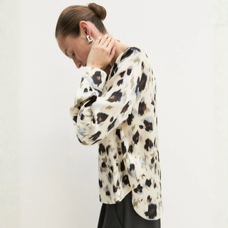 A woman wearing a long-sleeve blouse with an abstract black and gray pattern on a cream background, paired with black trousers, photographed against a plain off-white wall.