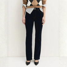 Load image into Gallery viewer, A model is shown from the chest down wearing dark navy blue straight-leg jeans, a brown and white argyle sweater vest, a striped shirt, and black pointed-toe heels against a white textured wall.
