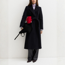 Load image into Gallery viewer, A model wearing a long navy  wool coat with a matching waist tie belt, styled with a purple turtleneck, black leather pants, and a black handbag with a bright red scarf accent, set against a clean white textured wall.
