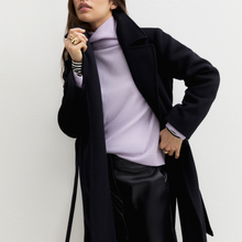 Load image into Gallery viewer, A woman wearing a long navy coat over a light purple turtleneck sweater and black pants, standing against a plain off-white wall.
