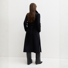 Load image into Gallery viewer, A woman viewed from behind wearing a long navy wrap-style coat with a waist tie, paired with black wide-leg boots, standing against a clean white studio background.
