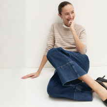 Load image into Gallery viewer, A woman sitting on a white floor against a white wall, wearing a beige and white striped knit sweater, wide-leg blue denim pants, and black pointed-toe heels.
