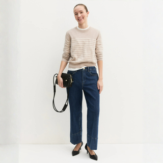 A woman stands against a plain white studio background wearing a beige and white horizontally striped knit sweater, dark blue wide-leg jeans, black pointed heels, and holding a small black crossbody bag.