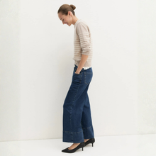 Load image into Gallery viewer, A woman stands in profile wearing a beige and white striped long-sleeve top, dark blue wide-leg cropped jeans with rolled hems, and black pointed-toe heels against a clean white studio background.
