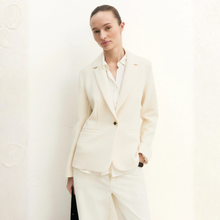 Load image into Gallery viewer, A woman wearing a tailored cream-colored blazer and matching trousers over a white collared shirt, standing against a light textured wall.
