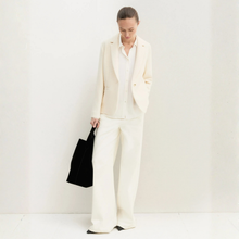 Load image into Gallery viewer, A woman stands against a plain white wall wearing a cream-colored tailored suit with a blazer, wide-leg trousers, and a white shirt. She is holding a black tote bag.
