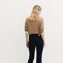 Load image into Gallery viewer, A woman with blonde hair is shown from the back wearing a tan crewneck sweater with striped cuffs and dark blue jeans, photographed against a plain white studio background.
