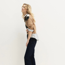 Load image into Gallery viewer, A woman posing in profile wearing a tan patchwork jacket over a white striped button-down shirt tucked into dark navy trousers, set against a plain off-white studio background.
