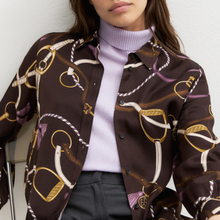 Load image into Gallery viewer, A close-up of a model wearing a dark brown button-up shirt with a gold, white, and purple equestrian-themed chain and bit pattern, layered over a light purple turtleneck sweater.
