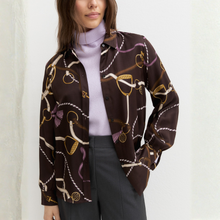 Load image into Gallery viewer, A woman wearing a dark brown long-sleeve button-up shirt with an equestrian-themed print of reins, bits, and bows in white, gold, and purple, layered over a light purple turtleneck sweater and dark gray trousers.
