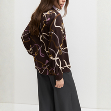 Load image into Gallery viewer, A woman wearing a dark brown long-sleeve blouse with a white, gold, and purple horsebit and chain pattern, paired with dark gray wide-leg trousers, shown from a side-back angle against a light gray wall.
