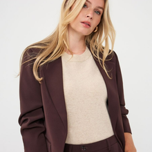 Load image into Gallery viewer, A blonde woman wearing a dark brown blazer over a light beige crewneck sweater, shown from the chest up against a plain light gray studio background.
