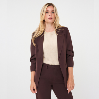 A blonde woman modeling a dark brown tailored blazer, matching trousers, and a cream knit top against a plain light gray studio background.