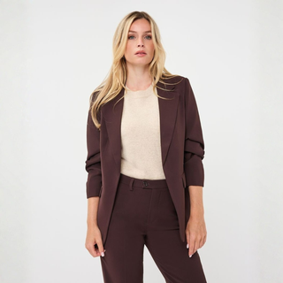 A blonde model wearing a dark brown tailored blazer and matching trousers over a cream knit top, standing against a light gray studio background.