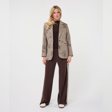Load image into Gallery viewer, A woman stands against a plain light gray studio background wearing a brown and beige patterned blazer over a dark brown turtleneck, matching wide-leg brown pants with white side stripes, and white shoes.
