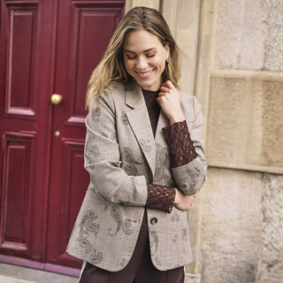 A woman wearing a beige and brown patterned blazer over a dark brown lace-sleeve top, standing outdoors in front of a red wooden door and a stone wall.
