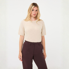 Load image into Gallery viewer, A blonde model wearing a beige short-sleeve knit top and dark brown trousers, standing against a plain white studio background.

