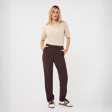 Load image into Gallery viewer, A woman modeling a beige short-sleeve knit top and dark brown straight-leg trousers with a matching belt, paired with white sneakers, against a plain light gray studio background.
