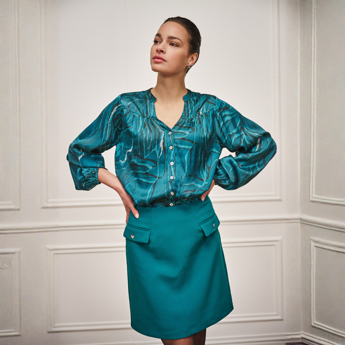 Woman wearing a teal patterned blouse and skirt against a white paneled wall.