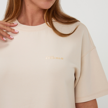 Load image into Gallery viewer, Beige t-shirt with gold logo worn by a person, on a plain background
