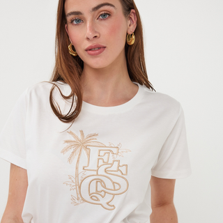 Woman wearing a white t-shirt with a gold logo and palm tree design on a plain background