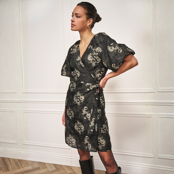 Woman wearing a black floral dress against a white paneled wall.