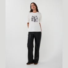 Load image into Gallery viewer, Woman wearing a white t-shirt with a graphic design and black pants on a white background
