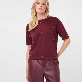 A woman modeling a short-sleeve crewneck knit top in deep burgundy with small white embroidered dots, paired with burgundy faux-leather pants, against a plain light gray studio background.