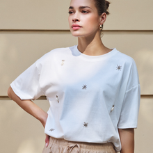 Load image into Gallery viewer, Woman wearing a white t-shirt with embroidered designs against a beige wall.
