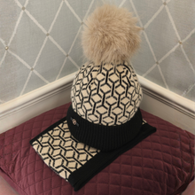 Load image into Gallery viewer, A knit beanie hat with a large beige faux-fur pom-pom, featuring a cream and black geometric pattern and a black ribbed cuff, displayed on a burgundy quilted cushion against a gray diamond-patterned wall.
