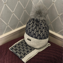 Load image into Gallery viewer, A stylish knitted beanie featuring a black and white geometric pattern, finished with a cosy white ribbed cuff and a fluffy grey faux-fur pom-pom. Displayed on a burgundy quilted cushion against a grey diamond-patterned background.
