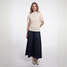 Load image into Gallery viewer, A woman modeling a beige short-sleeve knit top and a dark navy blue pleated maxi skirt, paired with beige ankle boots, against a plain light gray studio background.
