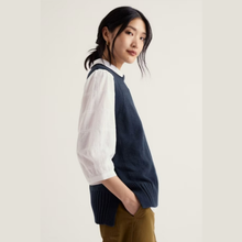 Load image into Gallery viewer, A woman wearing a navy blue knit sweater vest over a white textured long-sleeve shirt, paired with olive brown pants, photographed against a plain light beige studio background.
