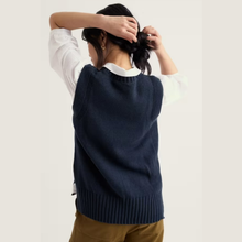 Load image into Gallery viewer, A woman shown from behind wearing a navy blue ribbed knit sweater vest over a white long-sleeve shirt and brown pants, with her hands in her hair against a plain light background.
