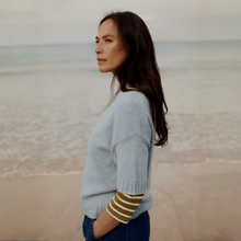 Load image into Gallery viewer, A woman with long dark hair stands in profile on a beach, wearing a light blue knit sweater with olive green and white striped elbow patches and dark blue jeans.
