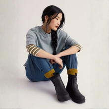 Load image into Gallery viewer, A woman sits on a light studio floor wearing a light gray knit sweater with striped cuffs, blue jeans with yellow ribbed cuffs, and black ankle boots with yellow lining.
