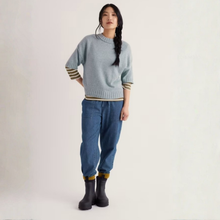 Load image into Gallery viewer, A model stands against a plain light background wearing a light blue knit sweater with striped cuffs and hem, blue jeans, and black boots.
