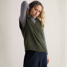Load image into Gallery viewer, A woman wearing an olive green knit sweater vest over a navy and white striped turtleneck, paired with navy trousers and a patterned headband, set against a plain off-white background.
