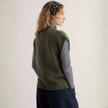 Load image into Gallery viewer, A woman shown from the back wearing an olive green knit sweater vest over a navy and white striped turtleneck sweater, paired with dark navy trousers and a patterned headband, against a plain off-white background.
