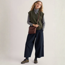 Load image into Gallery viewer, A woman stands against a plain light background wearing a dark olive green knit sweater vest over a black-and-white striped turtleneck, dark wide-leg pants, and brown shoes, with a small brown crossbody bag.
